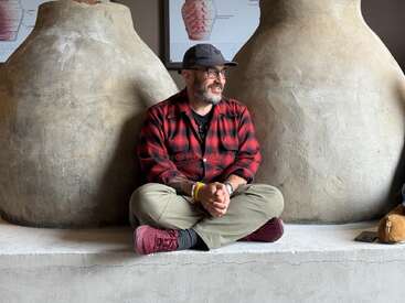 Um homem de camisa xadrez vermelha senta-se de pernas cruzadas, sorrindo, entre dois grandes vasos de barro. Ele usa óculos, boné e sapatos marrons. Dentro de casa.
