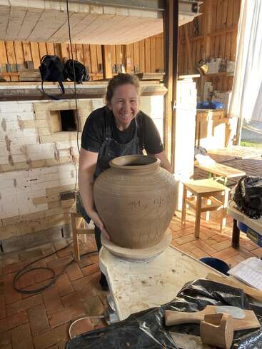 Uma mulher sorridente com um avental exibe orgulhosamente um grande pote de barro inacabado em um estúdio de cerâmica, cercado por ferramentas, móveis de madeira e um forno de tijolos.