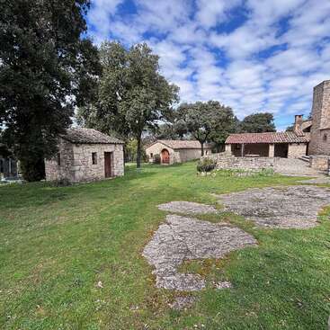 Esta imagem mostra uma cena rural tranquila com casas de pedra, grama verde, árvores e um céu azul nublado, evocando uma atmosfera rural rústica e pitoresca.
