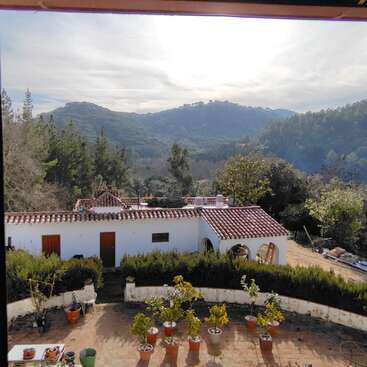 A imagem mostra uma casa branca serena com telhado de telha, cercada por uma vegetação exuberante e um pitoresco cenário montanhoso, exalando uma sensação de tranquilidade e beleza natural.