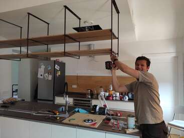 Um homem está em pé em uma cozinha, segurando uma furadeira elétrica acima de um balcão com ferramentas e uma pia, trabalhando em uma unidade de prateleiras.