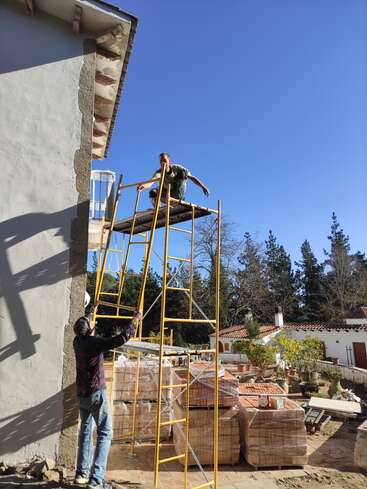 A imagem mostra dois homens trabalhando em um canteiro de obras, um deles em um andaime e o outro segurando uma escada, em um céu azul claro.