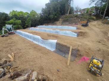 A imagem mostra um canteiro de obras com uma superfície de terra, com uma escavadeira branca, uma cerca de metal e um gerador amarelo, cercado por árvores e um céu nublado.