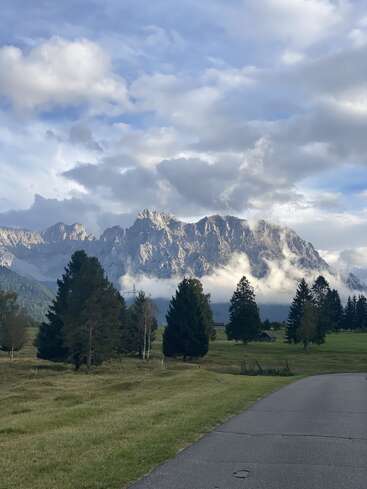 Eine malerische Berglandschaft mit dramatischen, wolkenbedeckten Gipfeln, grünen Wiesen, vereinzelten Kiefern und einem gewundenen, gepflasterten Weg, der sich unter einem strahlend blauen Himmel durch die friedliche Landschaft zieht.