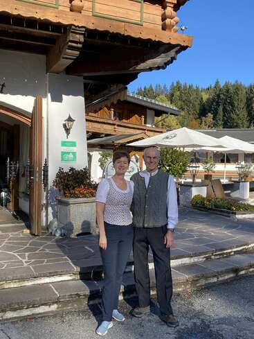 The image depicts a man and woman standing outside a building with a patio area, surrounded by trees and a blue sky. They are dressed in casual attire.