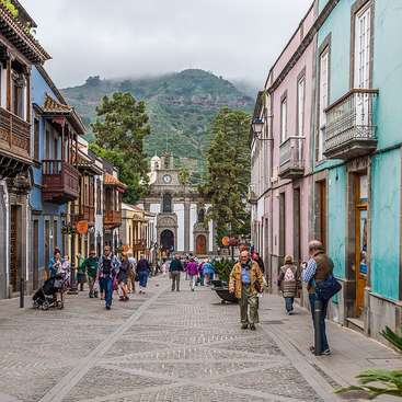 A imagem retrata uma charmosa rua de paralelepípedos em uma pequena cidade, repleta de edifícios coloridos e movimentada por pedestres, em um pitoresco cenário montanhoso.