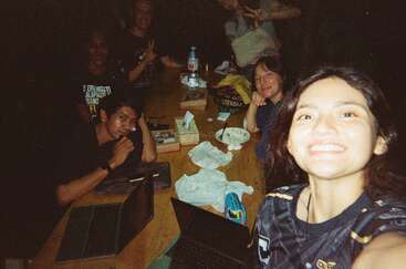 Eine Gruppe von Freunden versammelt sich nachts um einen Holztisch mit Laptops, Snacks, Getränken und einem Lächeln und macht ein fröhliches Selfie in einer gemütlichen, entspannten Atmosphäre.