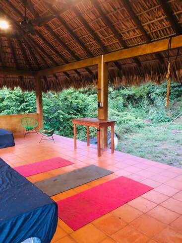 Cette photo montre un patio extérieur couvert d'un toit de chaume, trois tapis de yoga sur un sol carrelé, des chaises vertes, une table en bois et de la verdure luxuriante à l'extérieur.