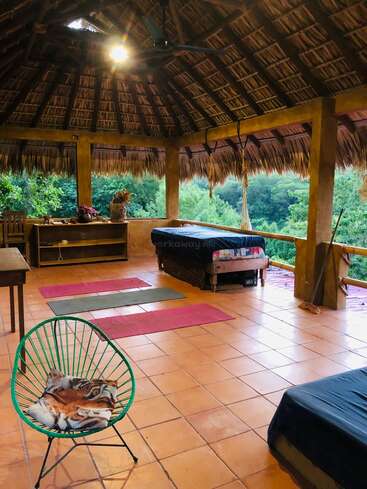 Une chambre en plein air avec un toit de chaume, un sol carrelé, des tapis de yoga, des lits, une chaise verte, des meubles en bois, entourée d'une végétation luxuriante et d'une vue naturelle paisible.