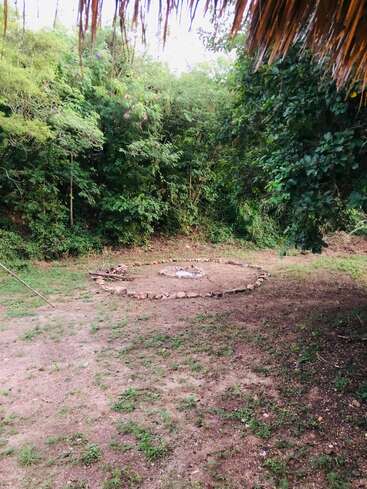 Cette image montre un foyer circulaire en pierre entouré de terre et d'herbe, situé dans une clairière à côté d'une forêt dense et verte, sous un toit de chaume.