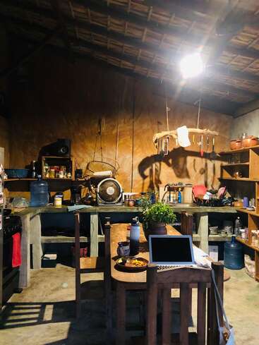 Une cuisine rustique avec des étagères ouvertes, une table en bois, un ordinateur portable, des plantes et des ustensiles suspendus au plafond. L'éclairage chaleureux crée une atmosphère douillette, habitée et accueillante.