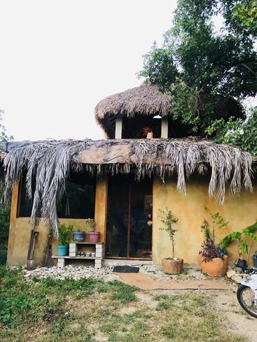 Cette image montre une petite maison rustique au toit de chaume, entourée de verdure. Des plantes en pot et un vélo sont visibles devant l'entrée.