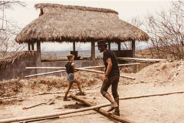 Two people work together carrying bamboo poles outside a rustic, thatched-roof hut in a dry, rural area, surrounded by scattered construction materials and barren trees.