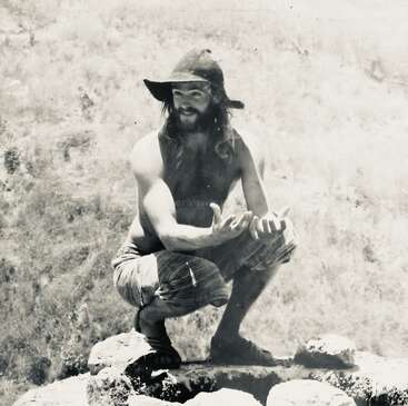 A bearded man squats on rocks outdoors, shirtless and wearing a wide-brimmed hat and striped shorts, smiling with outstretched hands, in a vintage black-and-white photograph.