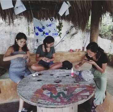 Three young women sit around a rustic, painted wooden table under a thatched roof, engaged in crafting activities. Colorful handprints hang on the wall behind them.
