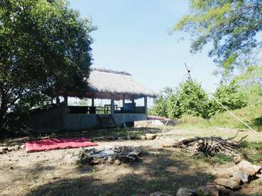 A rustic thatched-roof hut sits in a clearing surrounded by trees. There’s a fire pit, makeshift wood pile, red mat, and tripod structure outdoors. Peaceful setting.