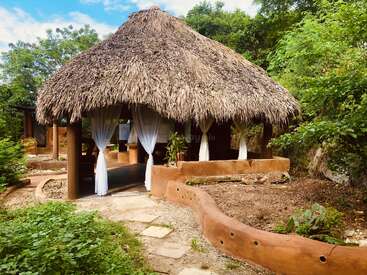 This image shows a rustic hut with a thatched roof and open sides, surrounded by lush greenery and a curved earthen pathway, evoking tranquility and nature.
