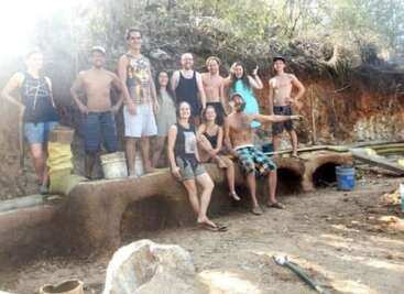 A group of eleven people pose outdoors on a curved, earthen bench. They appear happy, relaxed, and casually dressed, surrounded by trees and natural earth.