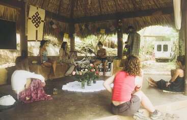 A group of people sit and relax in a rustic, open-air hut with a thatched roof. Flowers, decorations, and sunlight create a serene, communal atmosphere.