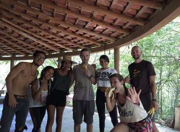 Seven friends pose cheerfully under a rustic wooden roof, some eating bananas. They appear happy and relaxed, enjoying time together in a bright, natural setting.
