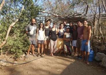 A group of twelve people stands outdoors in front of a rustic wooden structure, surrounded by trees and sunlight, smiling and appearing relaxed in casual clothing.