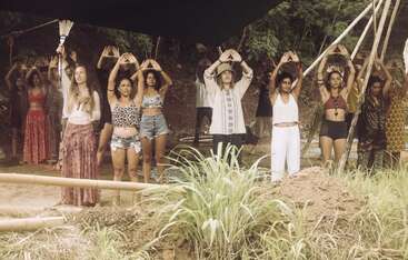 A group of people, dressed in bohemian-style clothing, stand outdoors in a circle, holding their hands above their heads in a triangular shape, participating in a ritual.