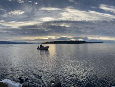 Ein kleines Boot mit zwei Personen schwimmt bei Sonnenuntergang auf einem ruhigen See. Der Himmel ist dramatisch, die Wolken und die entfernten baumbedeckten Ufer spiegeln sich wunderschön.