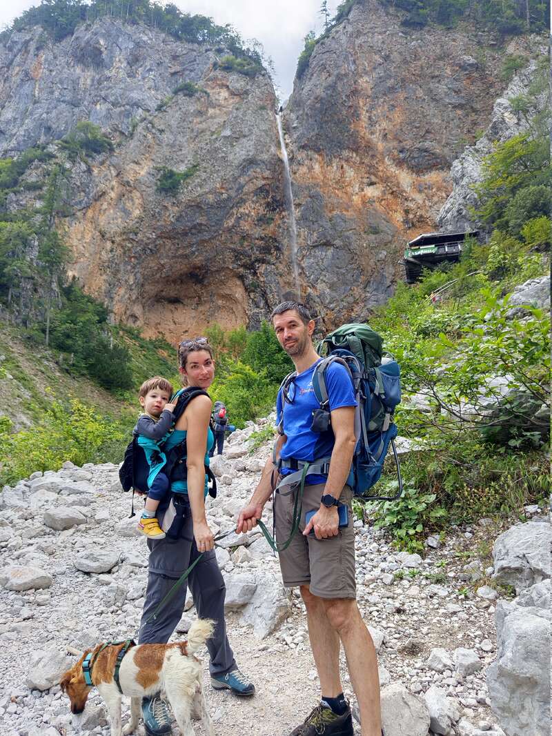 Eine Familie wandert auf einem felsigen Bergpfad. Die Mutter trägt ein Kind, der Vater hält einen Hund an der Leine. Sie sind umgeben von Grün und Felsen.