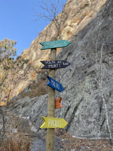 A wooden post with colorful directional signs points to destinations including "Mirante da Cruz," "Volta ao Monte," Malibu, and Paris, against a rocky outdoor backdrop.