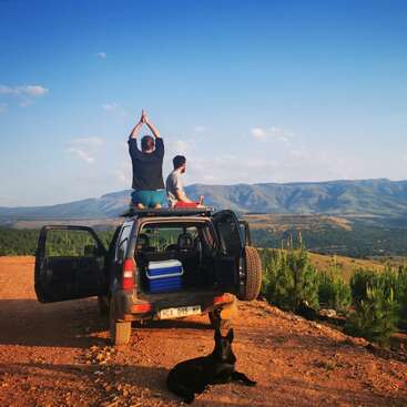 A man and woman sit on the roof of a 4x4 vehicle, overlooking a mountainous landscape, with a dog lying on the ground nearby.