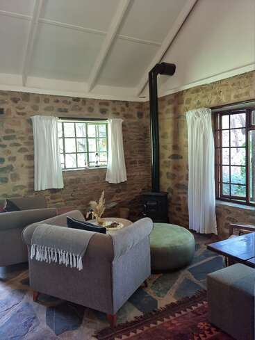 L'image représente un salon confortable avec des murs en pierre, un poêle à bois et un coin salon avec deux chaises grises et une table ronde.