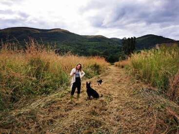 Une femme se tient dans un champ avec deux chiens, vêtue d'un cardigan blanc, d'un pantalon noir et de bottes vertes, tenant une tasse, avec des collines et des arbres en arrière-plan.
