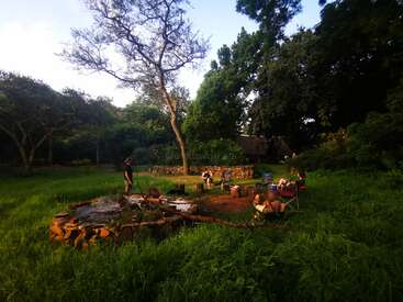 L'image représente un cadre extérieur serein avec un foyer, entouré de personnes, d'arbres et d'un mur de pierre, évoquant un sentiment de détente et de tranquillité.