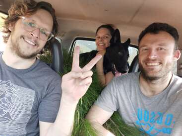L'image montre trois personnes et un chien à l'arrière d'une voiture, avec deux hommes au premier plan et une femme et un chien noir derrière eux.