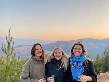 L'image représente trois femmes debout, chacune tenant un verre, avec un paysage montagneux et des arbres en arrière-plan, sur fond de soleil couchant.