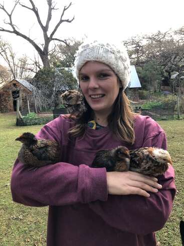 L'image représente une femme berçant trois poulets dans ses bras, sur un fond serein composé d'un champ herbeux et de bâtiments rustiques.