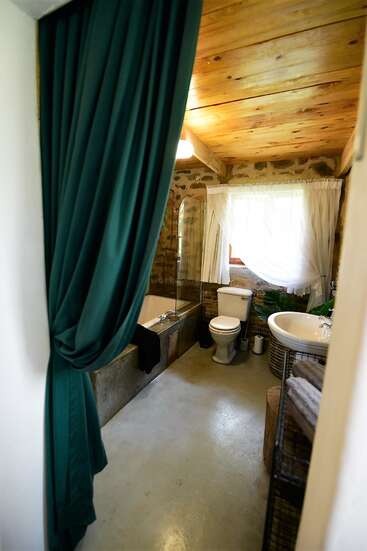 L'image représente une salle de bains rustique avec un plafond en bois, des murs en pierre, des toilettes, un lavabo et une baignoire blancs, avec un rideau vert et une fenêtre avec des rideaux blancs.