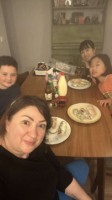 Four people, including two children and two adults, are smiling around a wooden table set with plates of food, condiments, and sauces. Cozy, homey atmosphere.