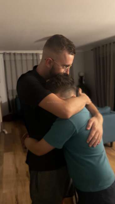 Two people share a heartfelt embrace in a warmly lit room. The taller person comforts the other, creating a strong sense of support, compassion, and friendship.