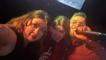 Three people are taking a selfie in a dimly lit room, happily holding microphones, possibly singing karaoke together, with warm lighting and joyful expressions on their faces.