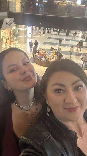 Two women smile for a selfie in a mall. Behind them, shoppers walk past stores and a large round display piled with colorful stuffed toys.