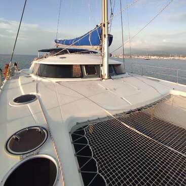 The image depicts a white catamaran sailboat with a blue sail cover, floating on the water, with a city skyline visible in the background.