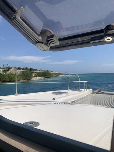 The image shows a view from a boat looking out toward a lush, green shoreline. The sea is blue, sky clear, and a hatch is open.