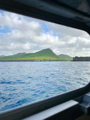 A scenic view from a boat window shows blue ocean waters, lush green hills, and a partly cloudy sky with sunlight gently illuminating the vibrant landscape outside.