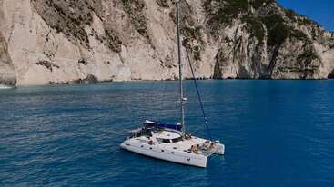 A white catamaran yacht floats serenely on crystal-clear blue water, with steep rocky cliffs in the background, under a bright sky, suggesting a tranquil getaway.