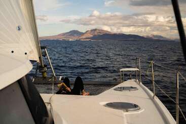 A person relaxes on the deck of a sailing boat, with calm blue ocean, distant mountains, and dramatic clouds creating a peaceful and scenic atmosphere.