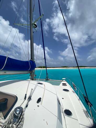 A white sailboat floats on crystal-clear turquoise water near a pristine sandy beach, with lush greenery and a vibrant blue sky filled with fluffy clouds above.