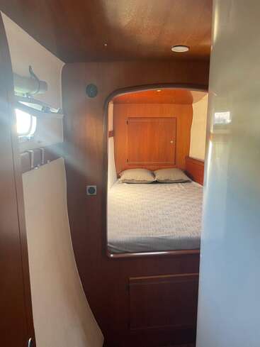 This image shows a cozy, compact bedroom with wood-paneled walls, a neatly made bed with two pillows, small shelves, and a porthole window bringing in light.