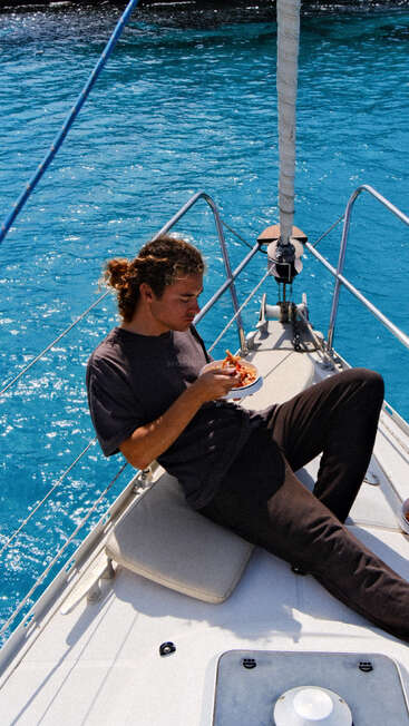 A person relaxes on the deck of a boat, enjoying pizza, surrounded by vibrant blue water under bright sunlight, creating a perfect moment of leisure.