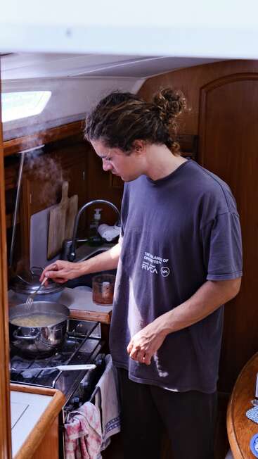 A person with long, curly hair tied back is cooking in a small boat kitchen, stirring a steaming pot on the stove with focused attention.
